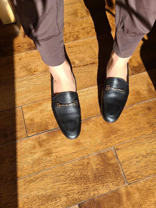 Person wearing black loafers with shiny buckles, standing on a wooden floor