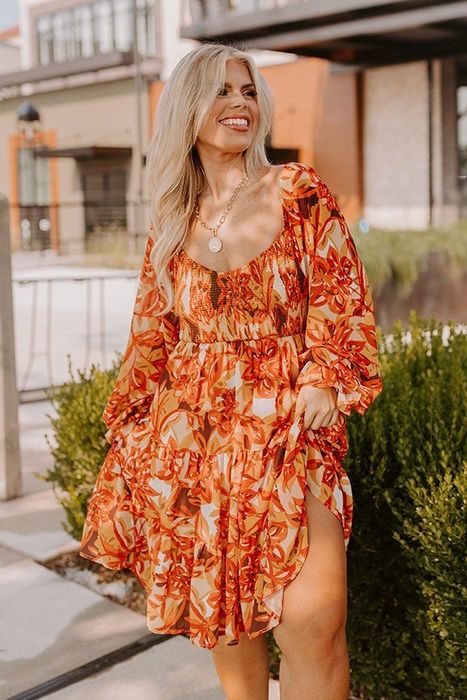 A model smiles outdoors while wearing a flowing floral mini dress with long sleeves and a high slit.