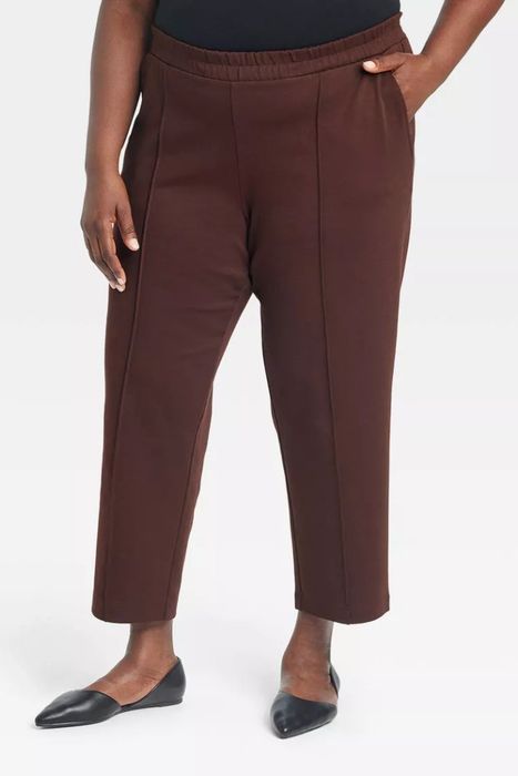 A person shown from the shoulders down, wearing dark, straight-leg trousers and black flats.