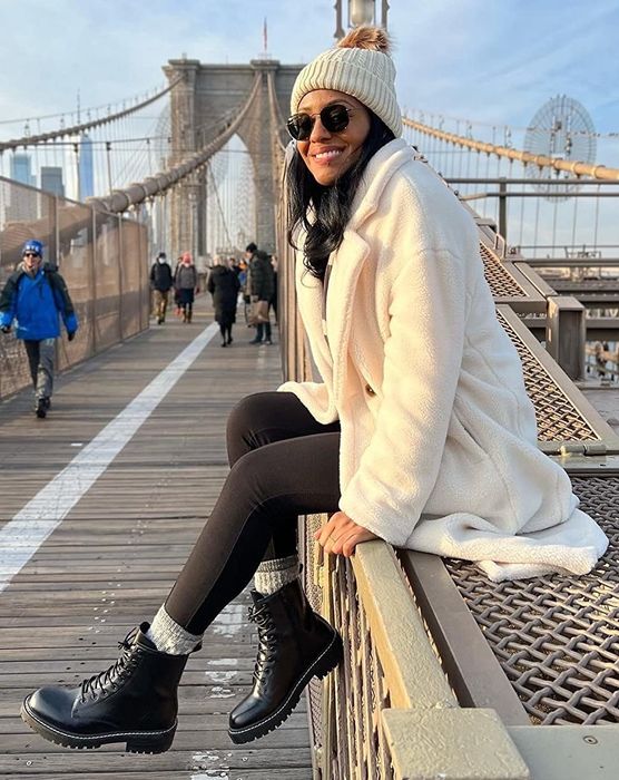 A reviewer wears the black boots paired with a white jacket, hat, and black pants while sitting on a bridge.