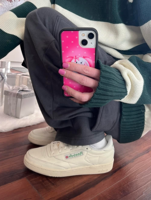 Person in a striped sweater with a phone case and sneakers crouches beside a white Christmas tree with presents underneath