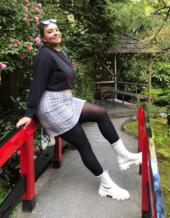 Another reviewer smiles while sitting on a red railing in a garden, dressed in the plaid skirt, black top, black tights, and white chunky boots.