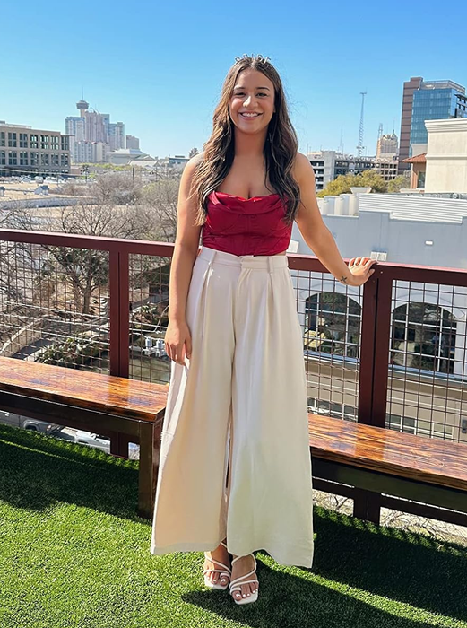 A reviewer dressed in a red strapless top and white wide-leg pants, standing on a balcony with a sweeping city view in the background.