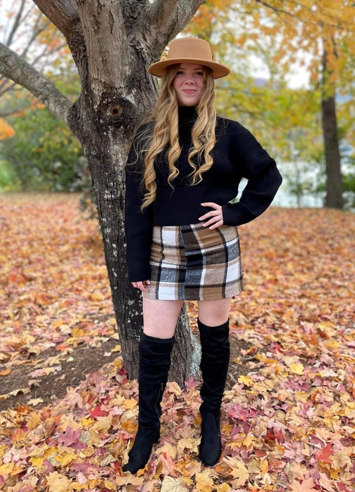 A reviewer stands outdoors in a plaid skirt, black turtleneck sweater, knee-high black boots, and a tan hat, surrounded by autumn leaves that have fallen around her.
