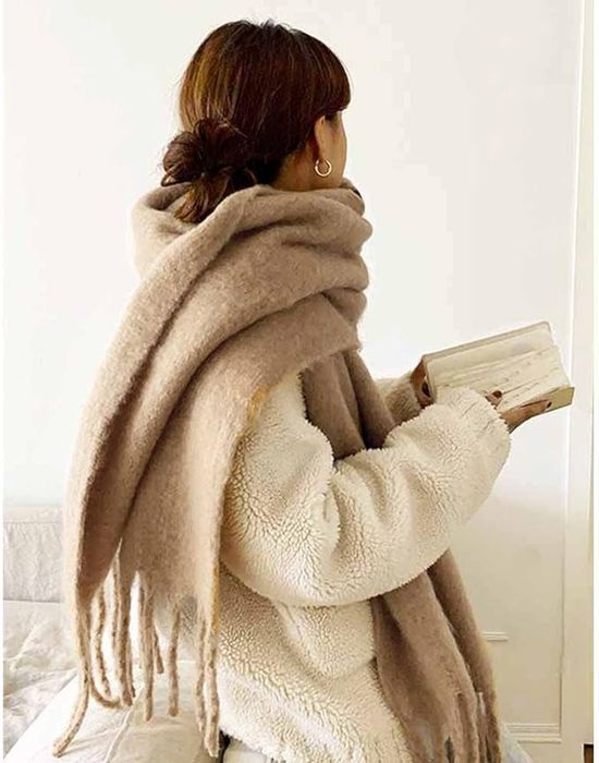 A model draped in a large camel-colored scarf with frayed edges.