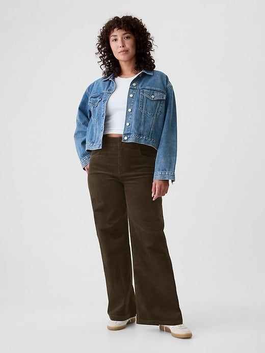 A model with curly hair dons a denim jacket over a white shirt and brown pants, posing against a plain background while gazing at the camera.
