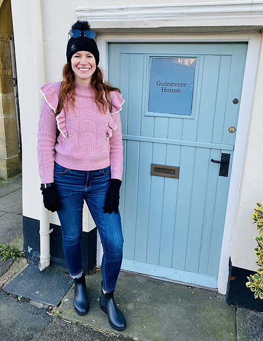Reviewer rocking black boots, paired with jeans and a pink sweater, standing next to a vibrant blue door.