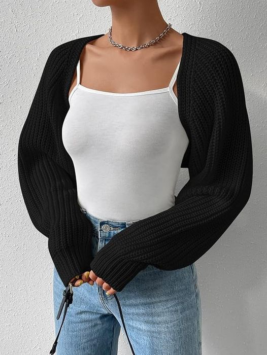 A model wears a white tank top, a black long-sleeve cardigan, and light blue jeans. A chain necklace completes the look, and their hands are partially visible.