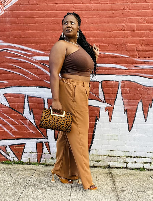 A reviewer strolling down the sidewalk, sporting a sleeveless top, high-waisted pants, and heels, holding a leopard print clutch. Urban mural setting in the background.