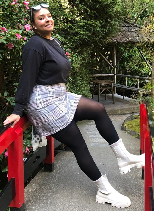 A reviewer poses on a small red bridge, surrounded by a lush garden, wearing a black top, plaid skirt, black tights, and white boots.