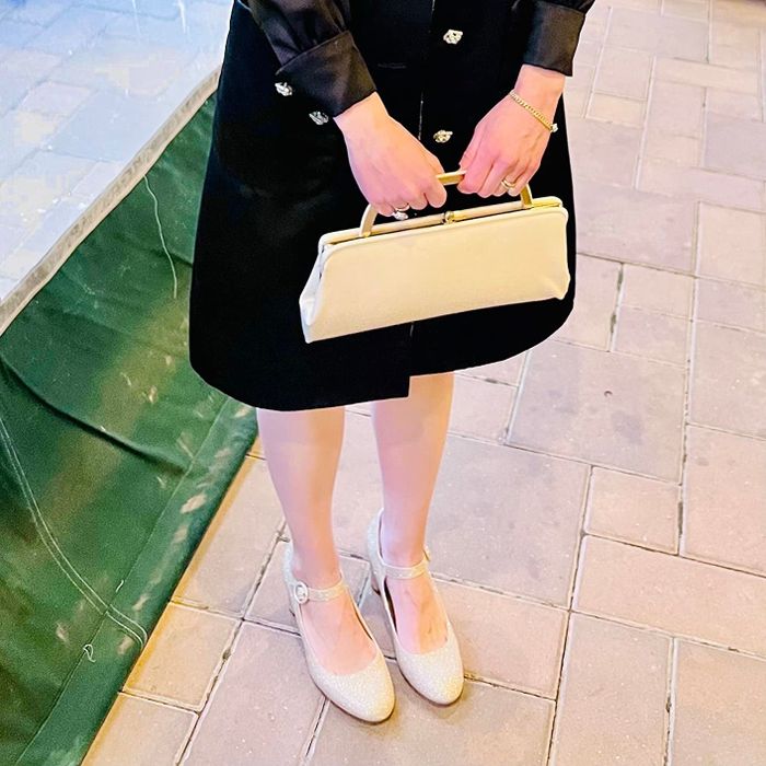 An individual dressed in an elegant ensemble, clutching a petite beige handbag, sporting ankle-strap heels, and positioned on a tiled surface.