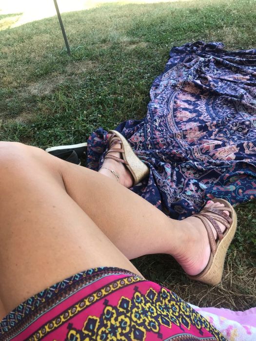 An individual seated on the grass wearing patterned shorts and wedge sandals, beside a vibrant, patterned blanket.