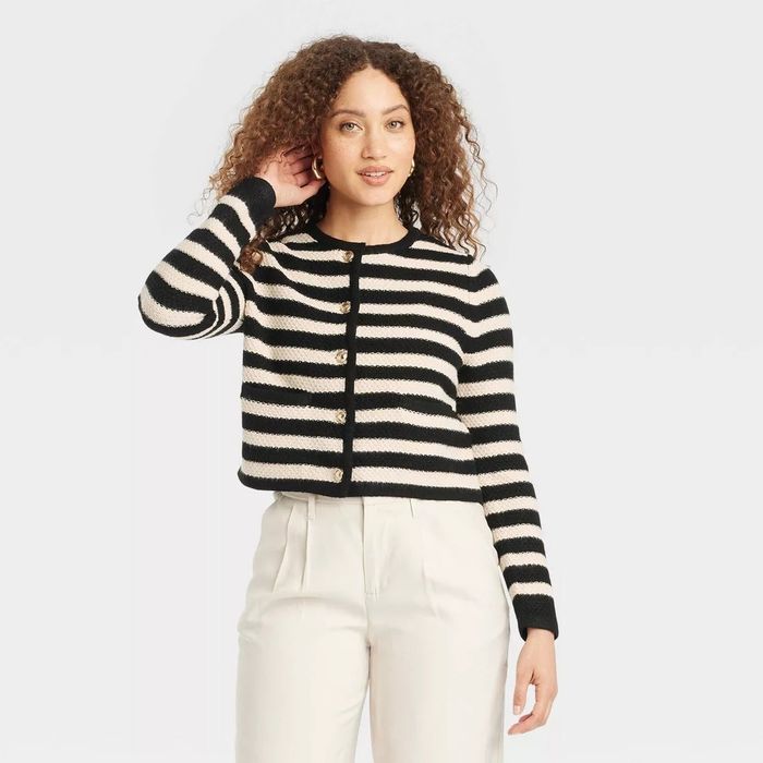 A person modeling a black-and-white striped button-up sweater with white pants, posing while gently touching her curly hair.