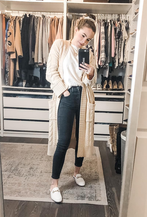 A reviewer snaps a mirror selfie in their walk-in closet, wearing a long cardigan, white top, black jeans, and white loafers. Various clothing items hang in the background.