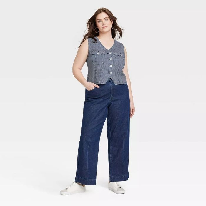A person exudes confidence while modeling wide-legged denim jeans paired with a sleeveless button-up denim vest. With hands casually tucked into the pockets, they complete the look with white sneakers.