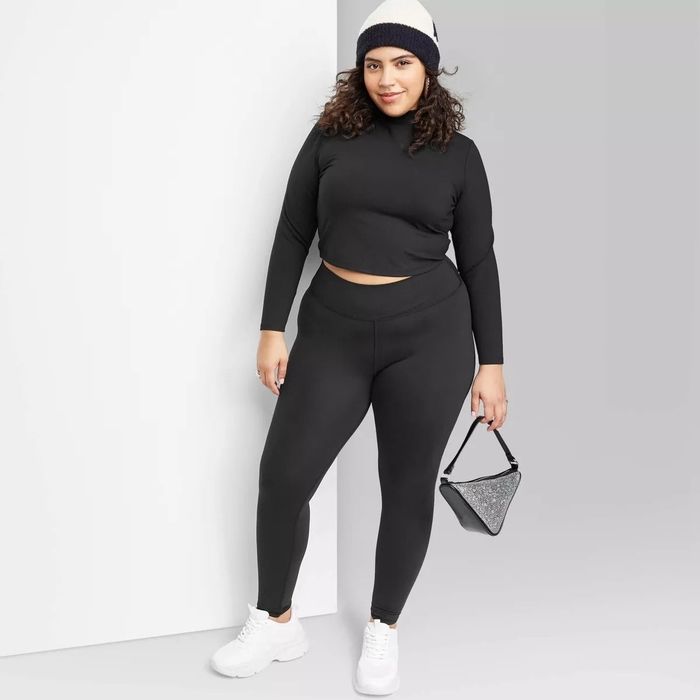 A person dressed in a sleek black long-sleeve crop top and leggings, carrying a small triangular handbag, wearing white sneakers and a beanie, striking a pose for a fashion article.