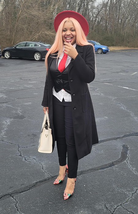 A reviewer exuding style in a black coat, striped tie, vest, and a red hat, smiling in a parking lot while holding a white handbag.