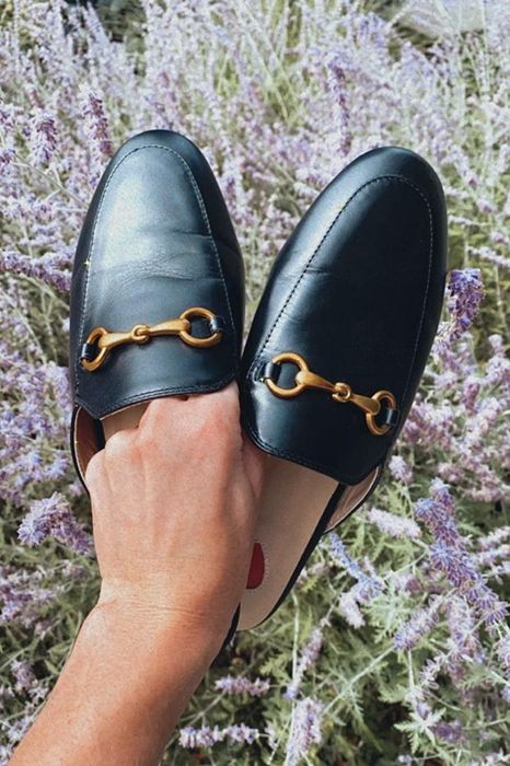 A person holds a pair of black loafer mules, showcasing the gold chain detail and open-back design.