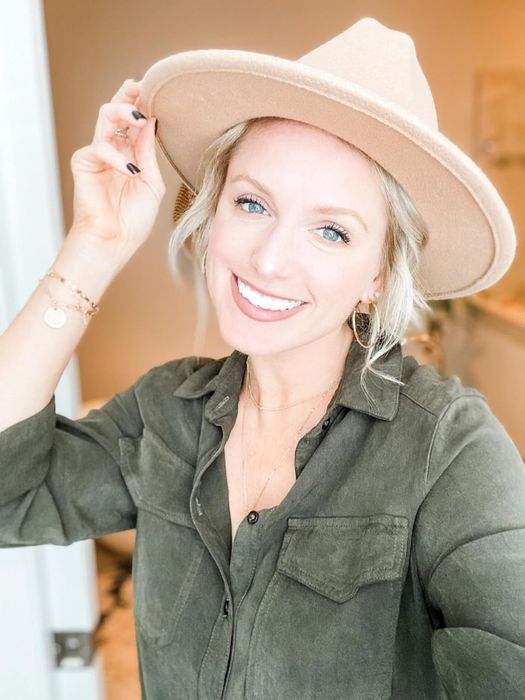 A reviewer smiles at the camera while holding her hat. She wears a button-up shirt and accessorizes with a bracelet and necklace.