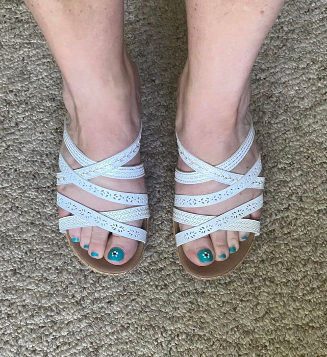 Feet adorned with elegantly crafted, strappy sandals on a carpeted floor, showcasing polished toenails.