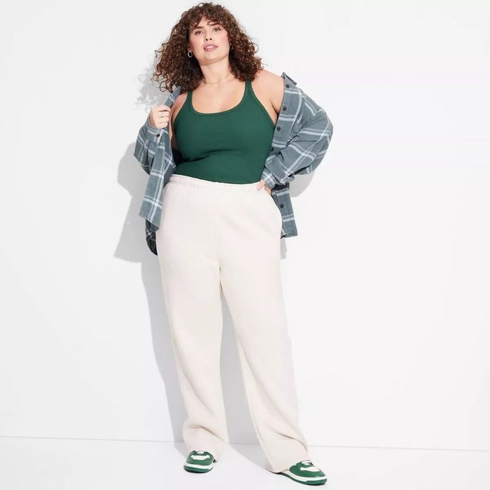 A person stands against a simple backdrop, wearing a green tank top, cream sweatpants, a gray plaid shirt, and green sneakers. Their curly hair complements the laid-back vibe.