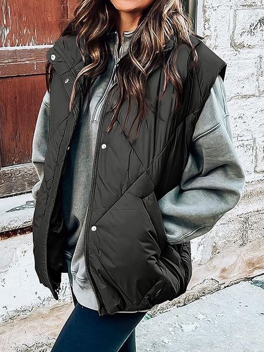 A model wears an oversized gray hoodie paired with a puffy black vest and dark pants, standing in front of a rustic stone wall.