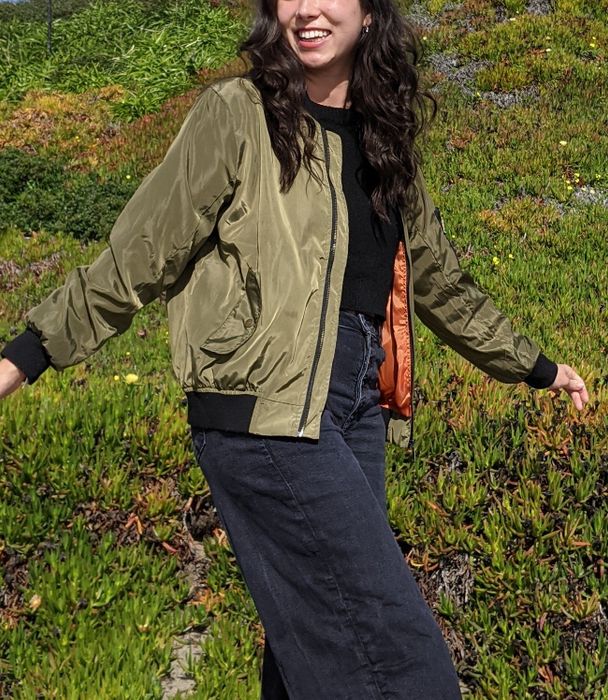 A reviewer with long, dark hair smiles as they walk outside. They’re wearing a green bomber jacket over a black shirt and dark pants.