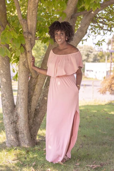 A reviewer poses outdoors in an off-shoulder, floor-length dress by a tree on a sunny day. The dress has a ruffled top layer and a side slit.