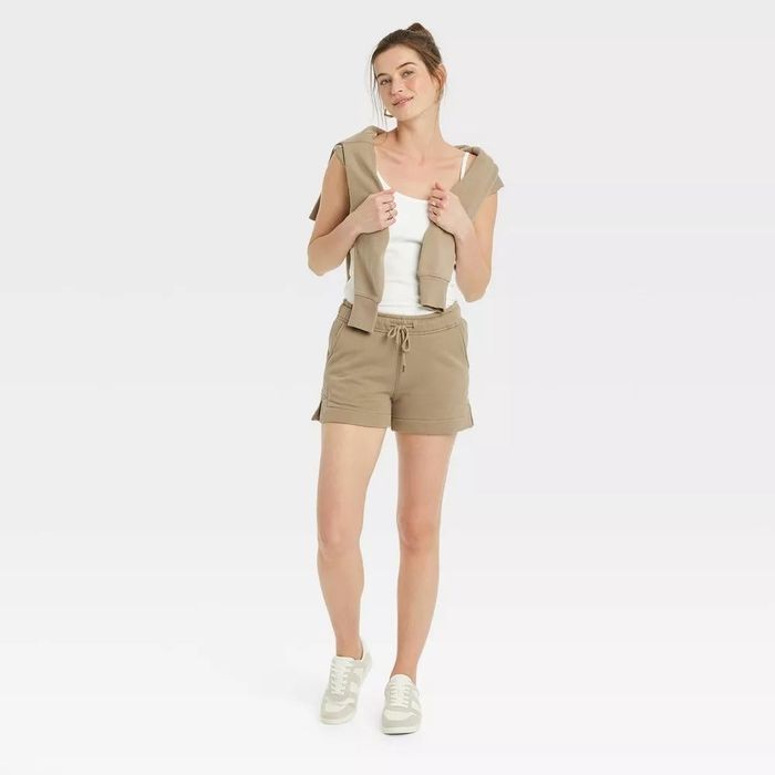 A model is dressed in a casual summer outfit featuring beige shorts, a white tank top, and a matching zip-up hoodie draped around her neck, paired with white sneakers.
