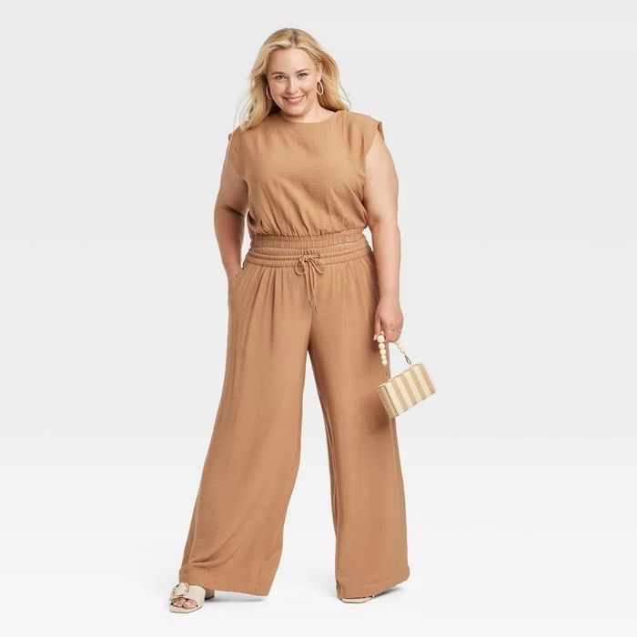 A person modeling a sleeveless jumpsuit with wide-leg pants and a belted waist, holding a small handbag.