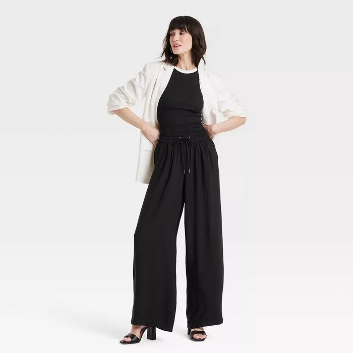 A person posing in a black top and wide-leg pants, paired with a white blazer. She completes the look with black high-heeled sandals and has one hand placed on her hip.
