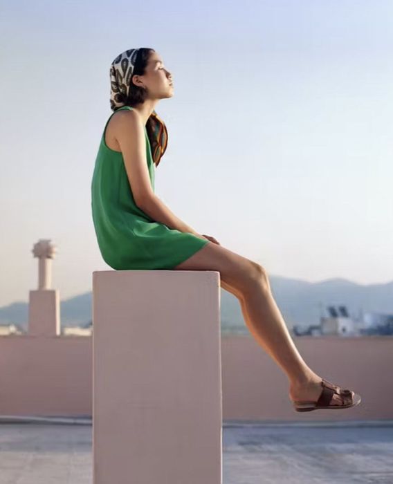 Model seated on a rooftop pillar, dressed in a green outfit paired with brown leather sandals