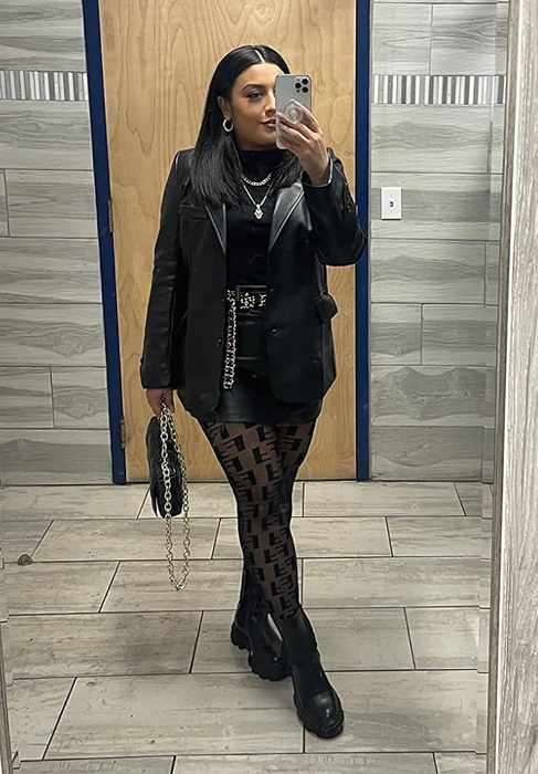 A reviewer strikes a pose in front of the mirror, showcasing a chic ensemble consisting of a black leather blazer, a black top, patterned tights, a black mini skirt, and bold accessories.