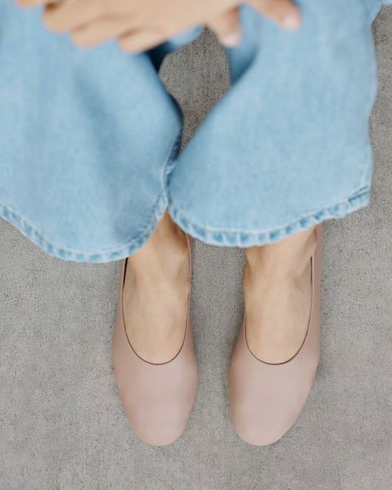 a close-up of a model wearing light-colored, minimalist flats and blue jeans, focusing on the shoes