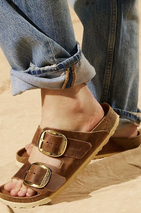 A model showcasing the dark brown sandals featuring large gold buckles, strolling on sandy terrain