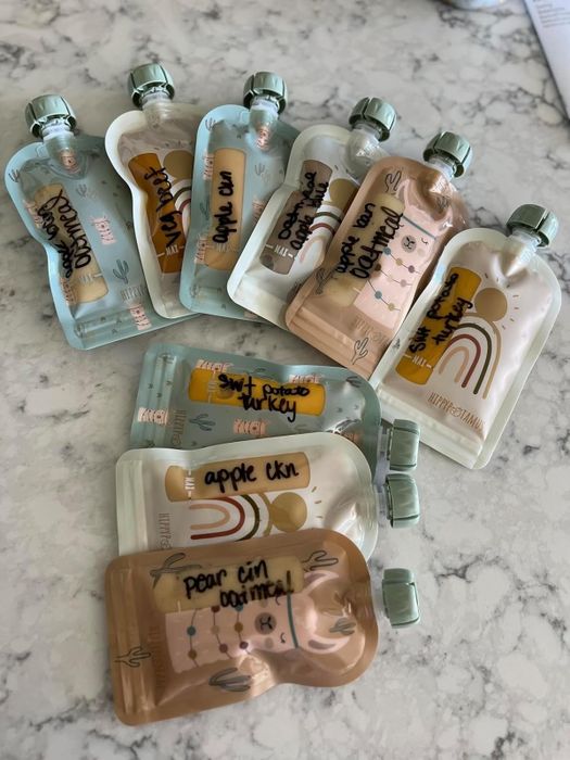 Nine baby food pouches with flavors like apple cinnamon, sweet potato, and pear, labeled in a handwritten style, displayed on a marble surface.