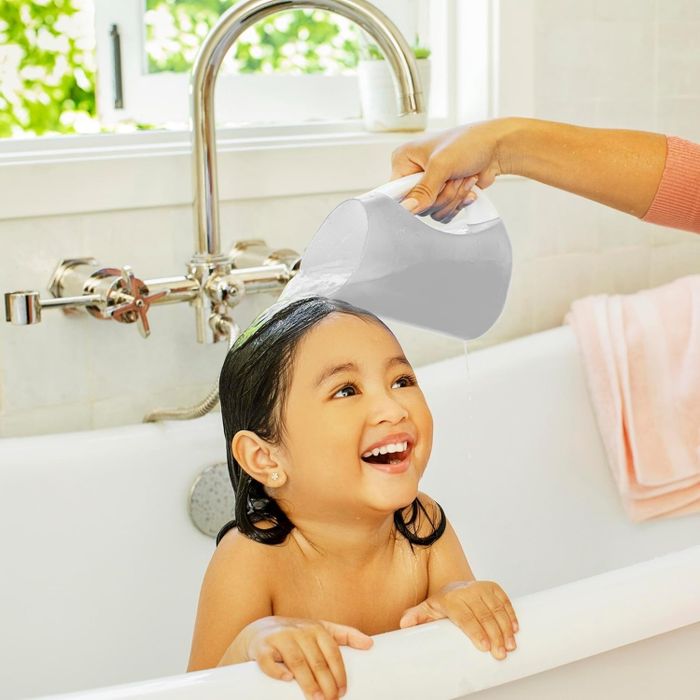 A toddler happily smiles in a bathtub while water is gently poured over their head using a rinsing cup.