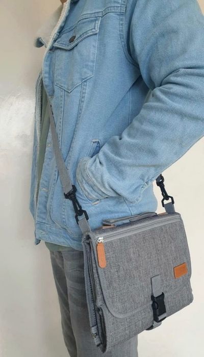A person wearing a denim jacket and gray pants, carrying a gray portable changing pad bag with a buckle as a crossbody.