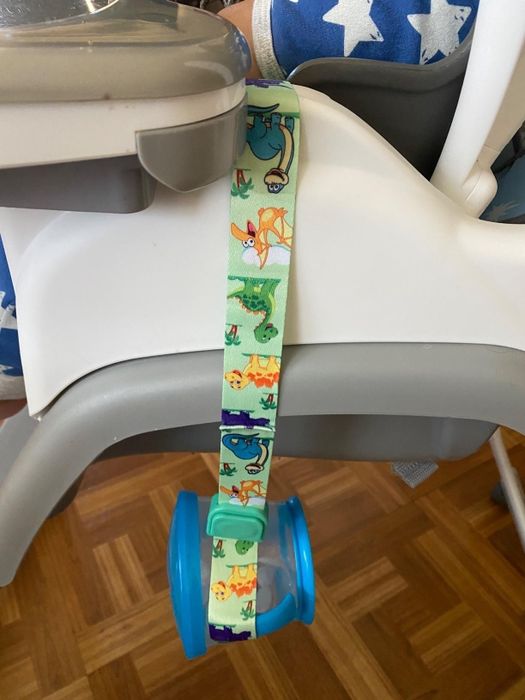 A snack cup secured with a vibrant dinosaur strap attached to a high chair, ensuring it stays put and doesn't tumble.