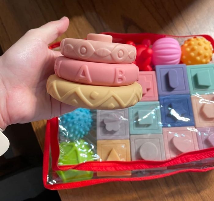 A person holding teething toys and textured blocks with geometric patterns, in front of a partially opened package on a wooden surface.