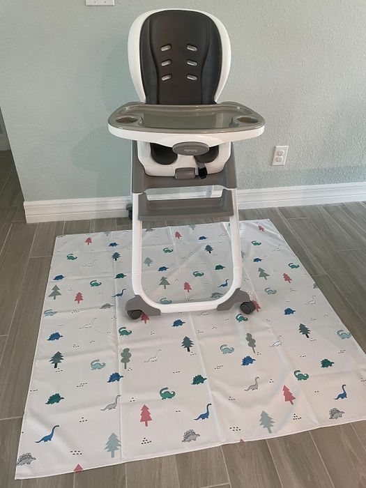 A high chair placed on a patterned mat featuring trees and animals, designed to protect floors during toddler meals. Perfect for home dining spaces.