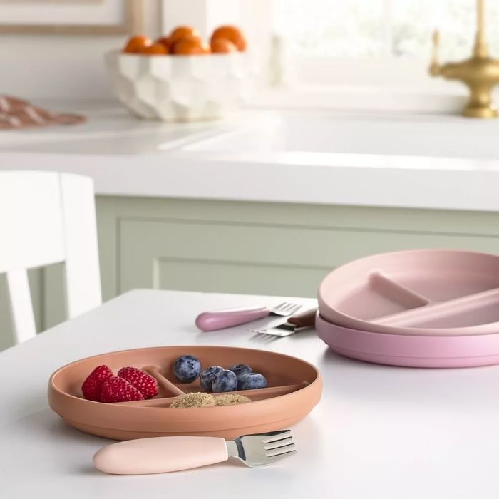 A set of divided silicone plates with fruit and snacks placed on a kitchen counter, with a stack of plates in the background.