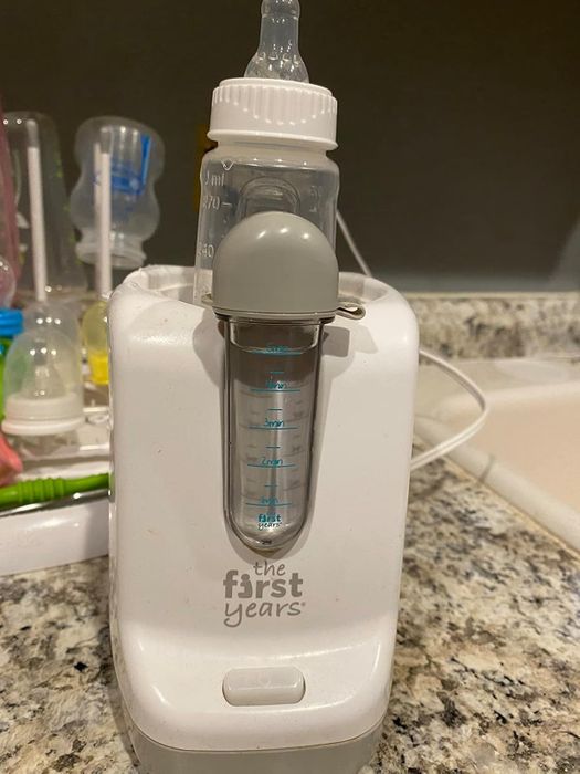 A baby bottle warmer on a kitchen counter with a bottle of milk inside. The brand name 'The First Years' is visible on the warmer.