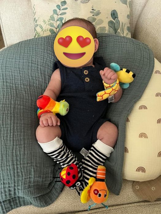 A baby sitting in a soft chair, playing with colorful wrist rattles and wearing striped socks.