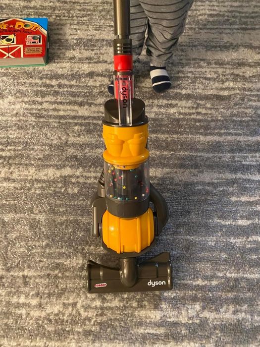 A child in striped pants is pushing a toy Dyson vacuum cleaner across a carpet.