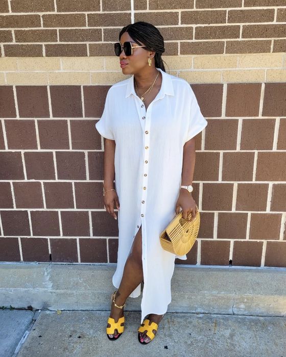 A reviewer in a white buttoned dress and yellow sandals, wearing sunglasses, strikes a pose by a brick wall while holding a fan-shaped handbag.