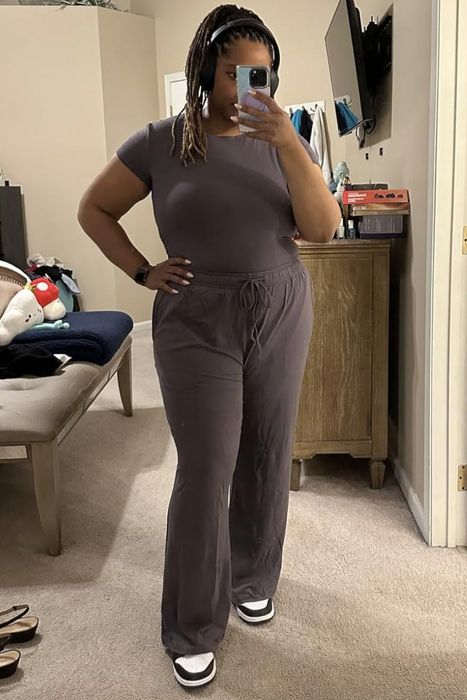 A woman poses for a mirror selfie in a bedroom, dressed in a casual short-sleeve top and loose-fitting pants. The room includes a couch with plush toys and a dresser with various items on top.
