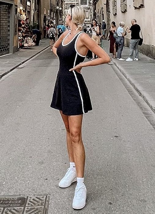 A person wearing a sleeveless black dress and white sneakers strikes a pose on a city street.