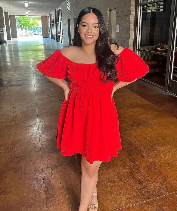 A reviewer poses in a hallway, wearing a red off-the-shoulder dress, smiling with hands placed on her hips.