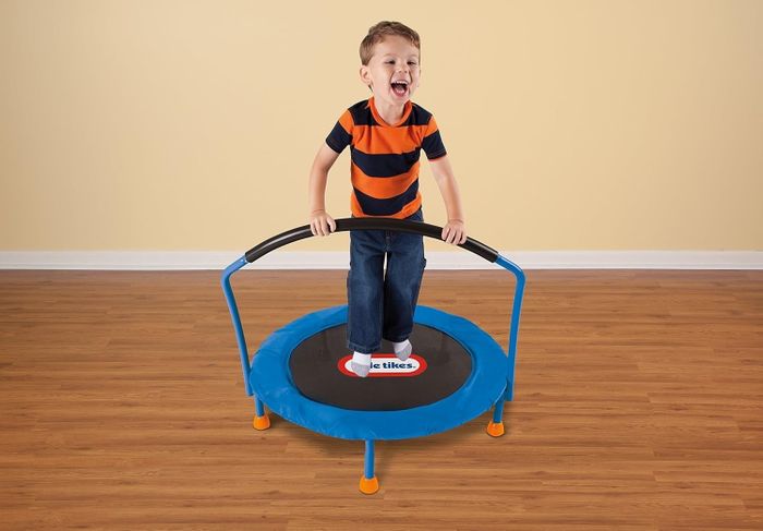 A young child in a striped t-shirt and jeans standing on a small trampoline, gripping a safety handlebar